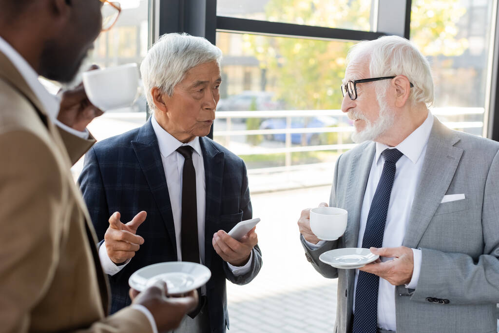 starszy azjatycki biznesmen ze smartfonem wskazując palcem podczas rozmowy z kolegami pijących kawę - Zdjęcie, obraz