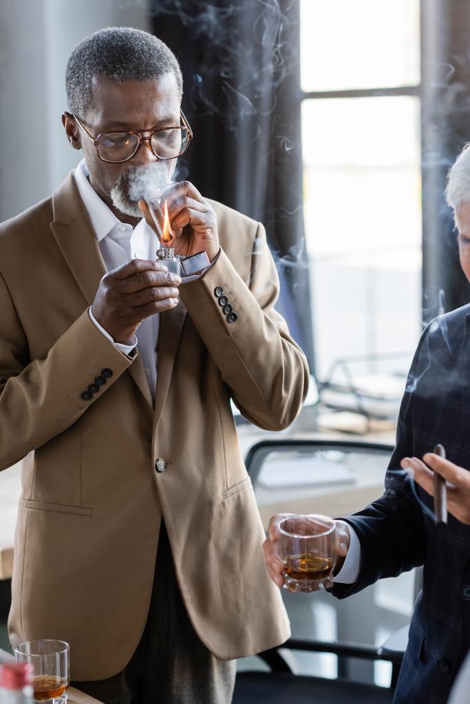 african american businessman in eyeglasses lighting cigar near colleague with glass of whiskey - Photo, Image