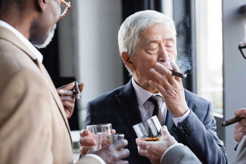 starszy azjatycki biznesmen palenie cygaro podczas brzęczenie szklanki whisky z zamazane międzyrasowych kolegów - Zdjęcie, obraz