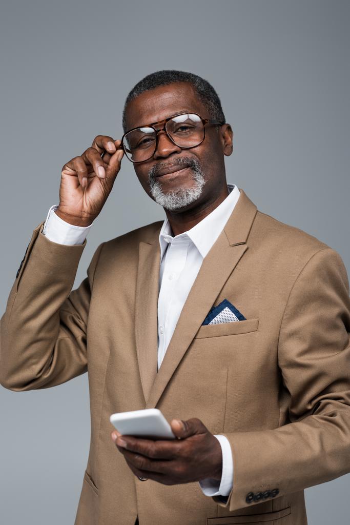 senior africký americký podnikatel s smartphone nastavení brýle při pohledu na kameru izolované na šedé - Fotografie, Obrázek