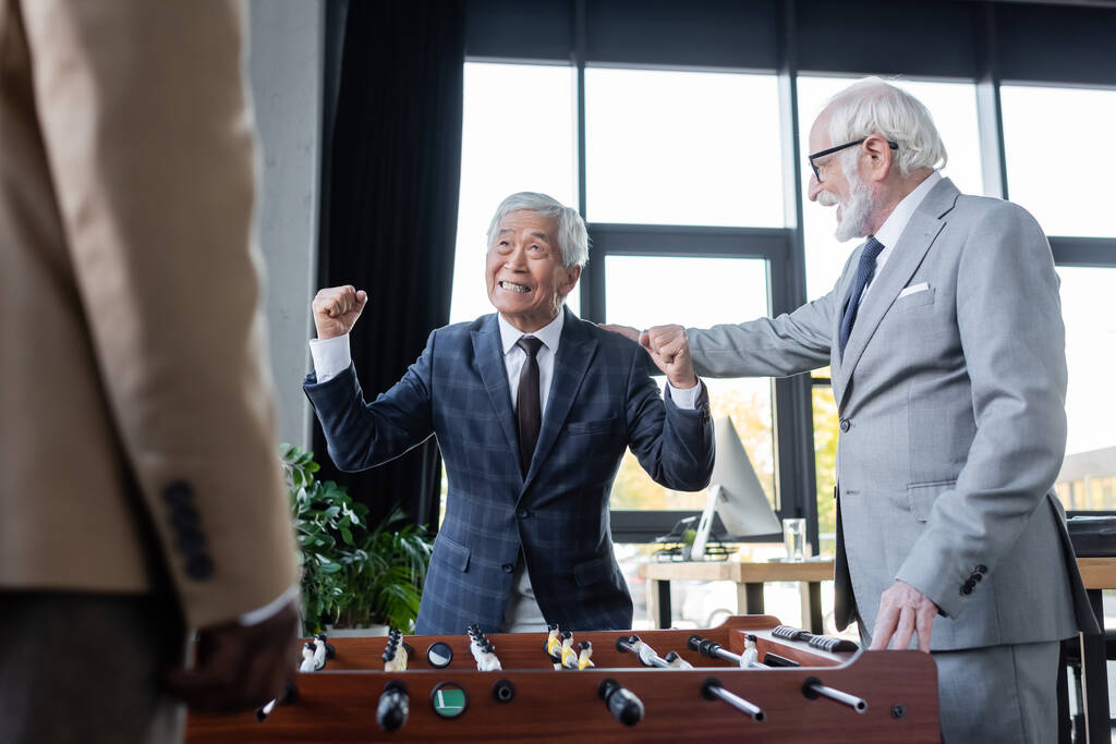 щасливий азіатський бізнесмен показує жест виграшу біля настільного футболу та старших багатоетнічних колег
 - Фото, зображення