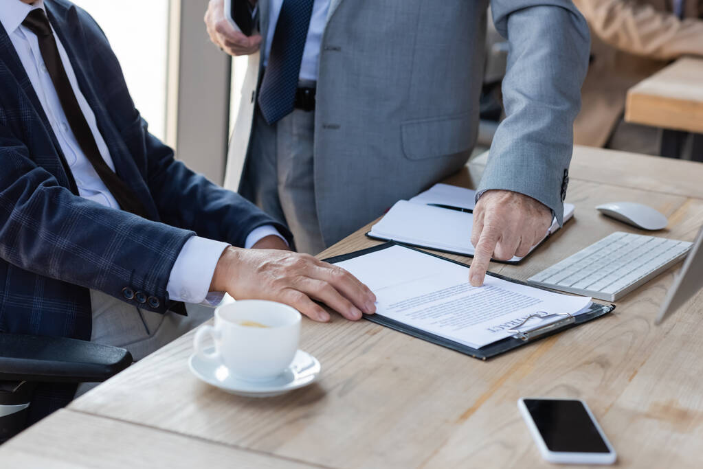 обрезанный взгляд старшего бизнесмена указывая на контракт рядом с коллегой и размытый кофе чашку - Фото, изображение