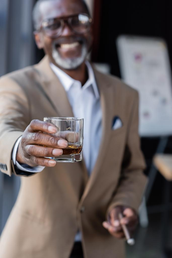 θολή αφροαμερικανός επιχειρηματίας χαμογελώντας κρατώντας ένα ποτήρι ουίσκι σε τεντωμένο χέρι - Φωτογραφία, εικόνα