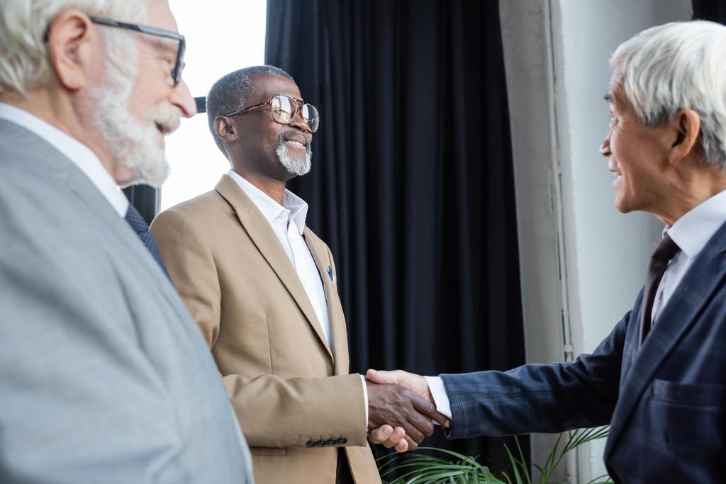 размытый старший бизнесмен улыбается рядом с африканскими американскими и азиатскими деловыми партнерами пожимая друг другу руки в офисе - Фото, изображение