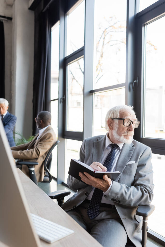 anziano uomo d'affari che scrive nel taccuino mentre guarda attraverso la finestra in ufficio - Foto, immagini
