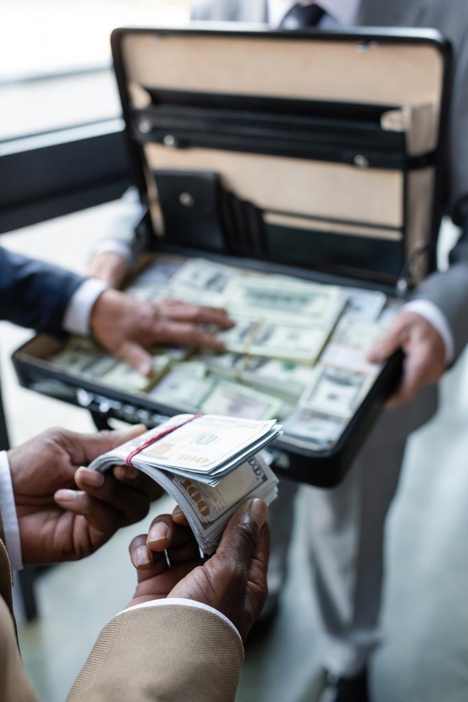 καλλιεργημένη άποψη του Αφροαμερικανού επιχειρηματία κρατώντας δολάρια κοντά θολή συναδέλφους και χαρτοφύλακα με χρήματα - Φωτογραφία, εικόνα