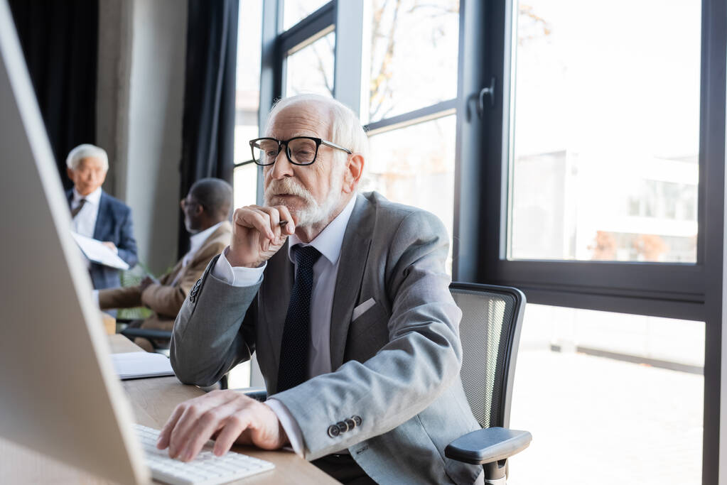 uomo d'affari anziano in occhiali che lavora su computer vicino a colleghi multiculturali offuscati - Foto, immagini