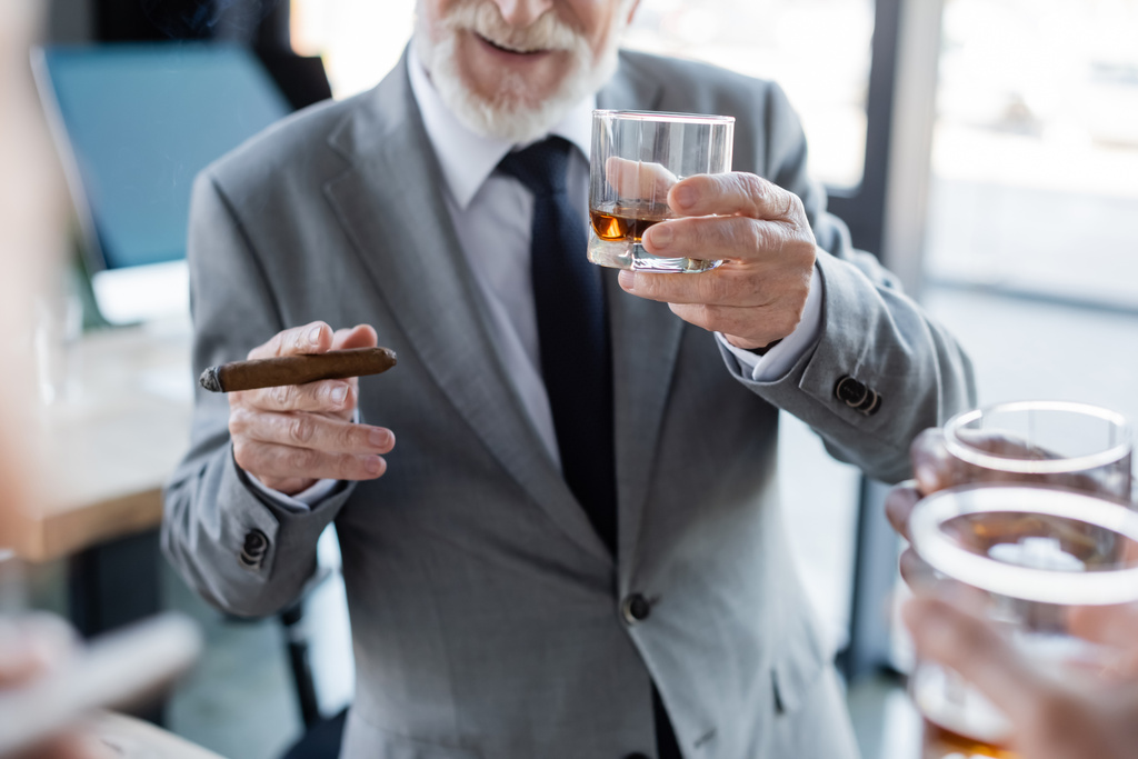 vista recortada del anciano hombre de negocios sosteniendo cigarro y vaso de whisky cerca de colegas borrosos - Foto, imagen