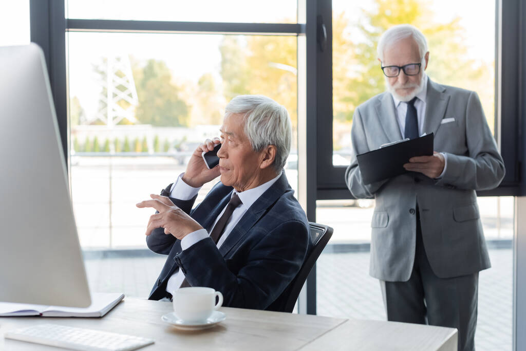 старший азіатський бізнесмен розмовляє на смартфоні біля комп'ютера і колеги з буфера
 - Фото, зображення