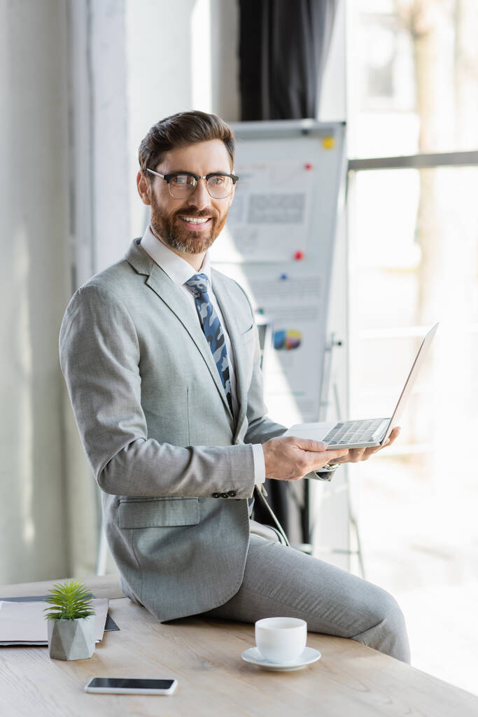 Mosolygós üzletember öltönyben gazdaság laptop közelében okostelefon és kávé az irodában  - Fotó, kép