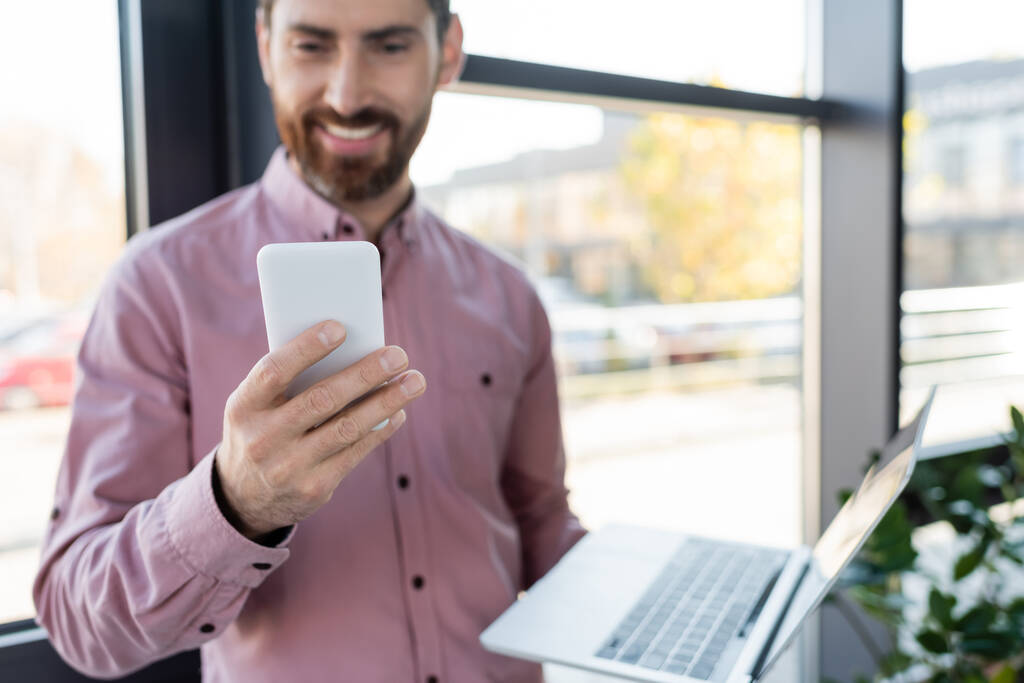 Smartphone v ruce rozmazaný podnikatel s notebookem v kanceláři  - Fotografie, Obrázek