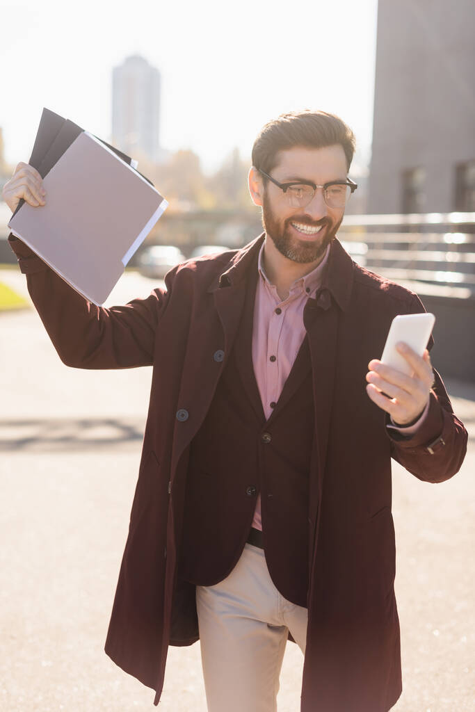 Homme d'affaires heureux tenant des dossiers en papier et utilisant un smartphone sur la rue urbaine  - Photo, image