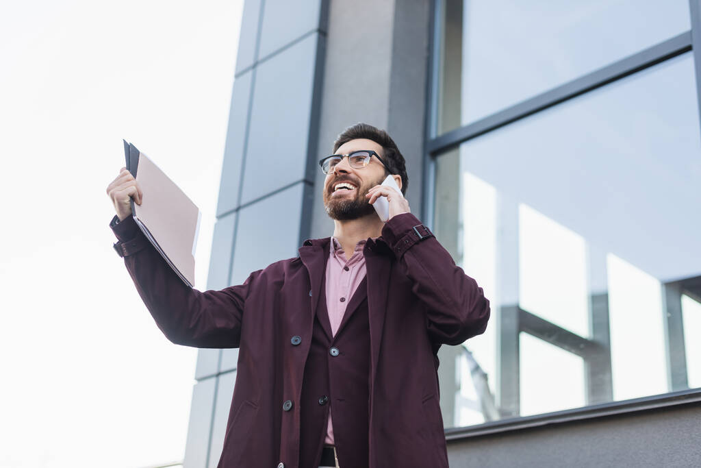 Низкий угол обзора веселого бизнесмена в плаще разговаривающего по смартфону и держащего бумажную папку возле здания на открытом воздухе  - Фото, изображение