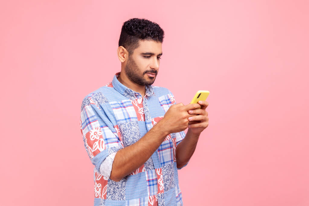 Side view of serious concentrated brunette man with beard holding cell phone in hands, chatting or checking e-mail, gadget addiction. Indoor studio shot isolated on pink background. - Photo, Image