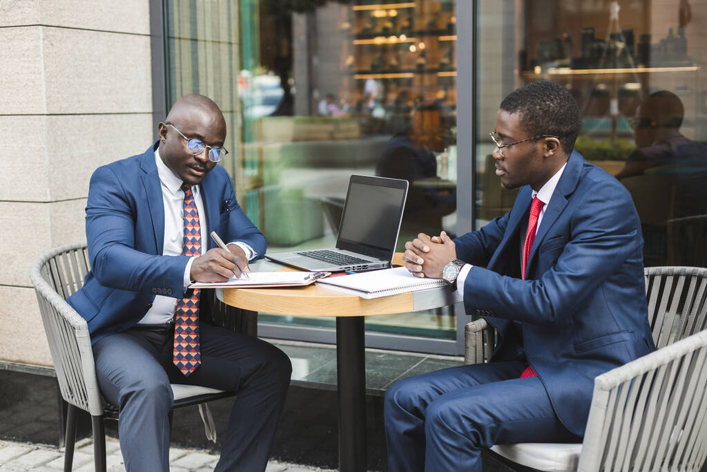 Twee donkergekleurde Afro-Amerikaanse zakenmannen in pakken en glazen met aktetassen zitten aan een tafel in een café in een buitenstad. - Foto, afbeelding