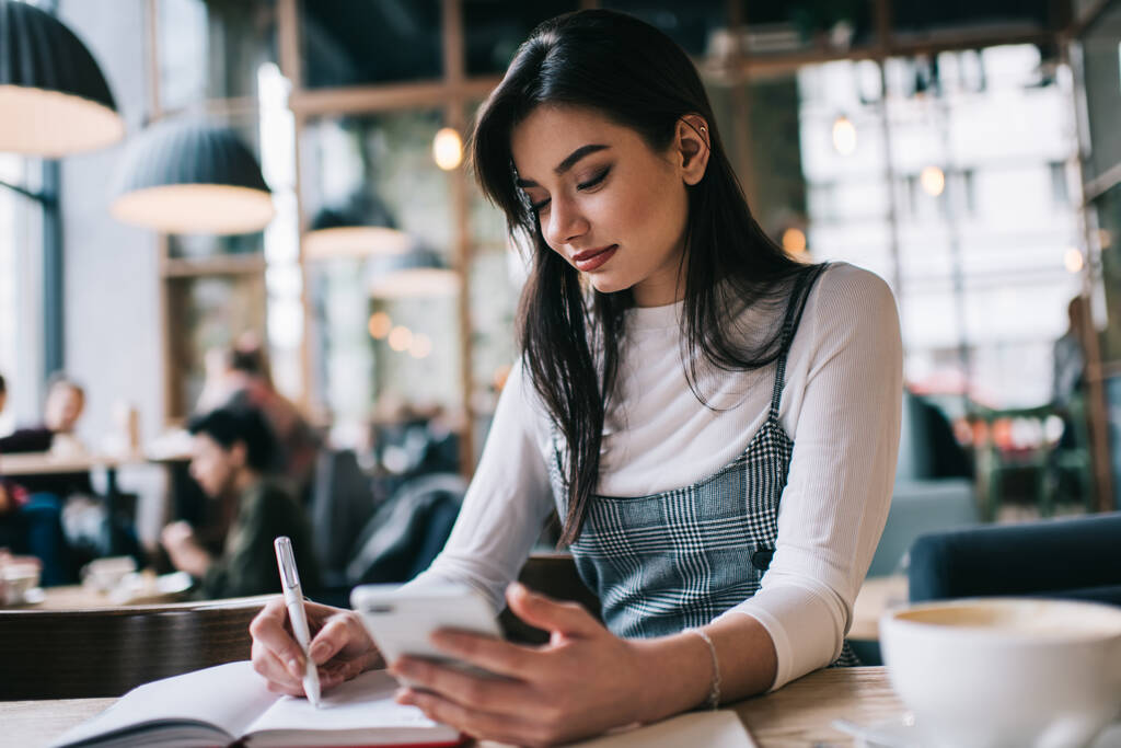 Увлекательная студентка, пользующаяся мобильным телефоном и записывающая заметки в блокнот, сидя за столом в светлом кафе и выполняя задание - Фото, изображение