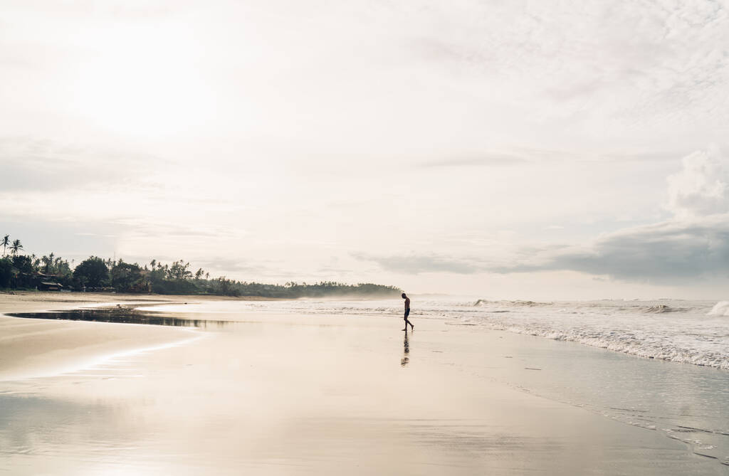 Боковой вид на неузнаваемого человека, идущего по мокрому песчаному берегу, рядом машущего морем под ярким сияющим солнцем и наслаждающегося гармонией - Фото, изображение