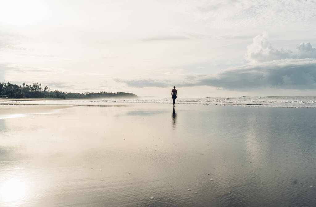 Vista trasera del hombre anónimo caminando lejos en la costa húmeda vacía cerca del mar bajo el sol brillante y disfrutando de estar solo - Foto, imagen