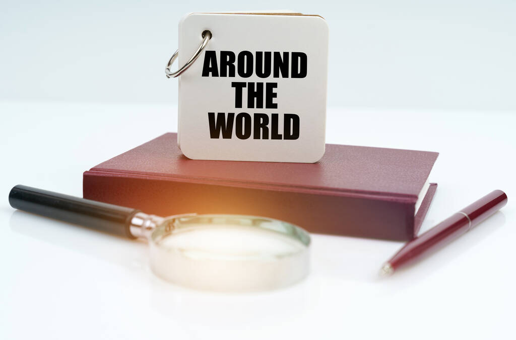 Em uma superfície branca encontra-se um caderno vermelho, uma caneta, uma lupa e um caderno com a inscrição - Around the World - Foto, Imagem