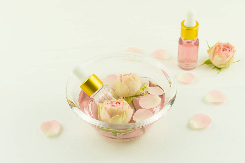 skleněná mísa s růžovou vodou, květy a okvětními lístky růží, kosmetické láhve s květinovým olejem zfir. aromaterapie, antistresová - Fotografie, Obrázek