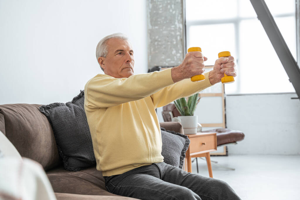 Старик делает упражнения с гантелями, сидя на диване и чувствуя себя серьезным - Фото, изображение