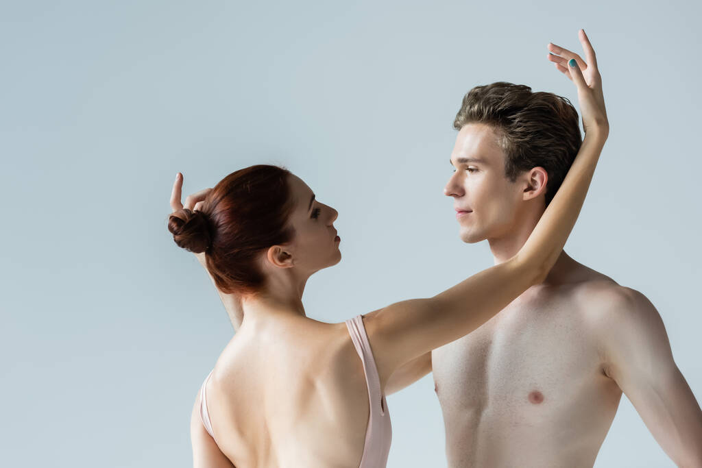 балерина смотрит на партнера без рубашки, исполняя танец, изолированный от серого  - Фото, изображение