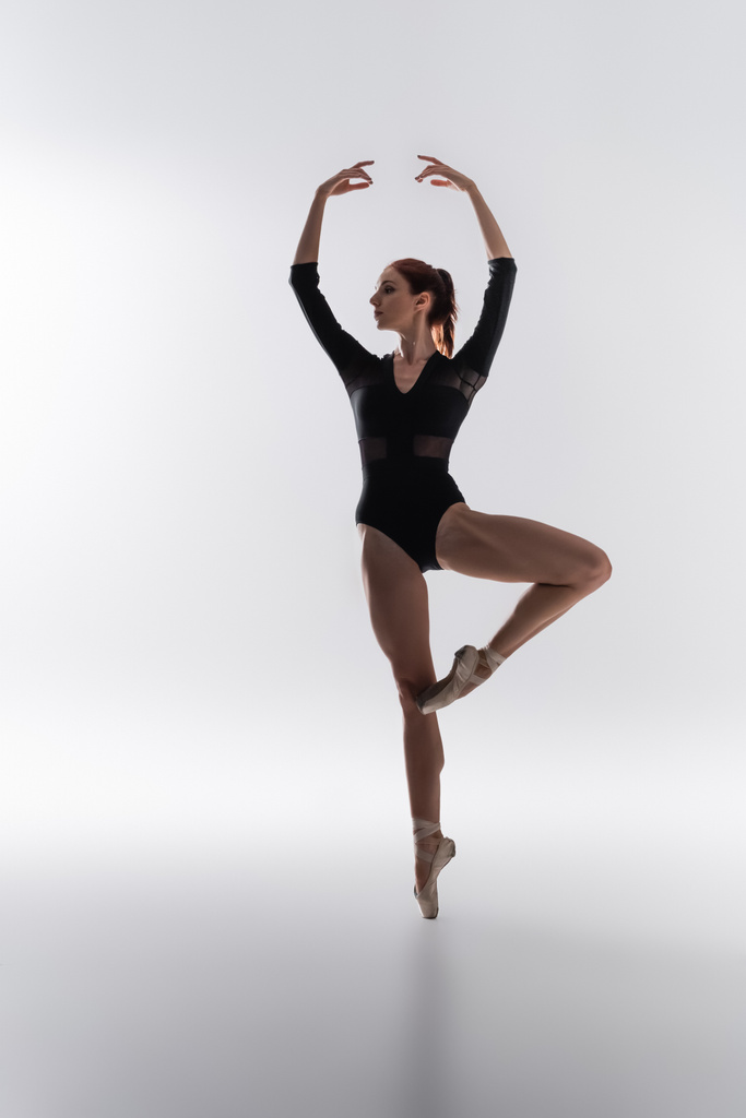 повна довжина елегантної балерини в боді-костюмі, що виконує танець з піднятими руками на сірому
 - Фото, зображення