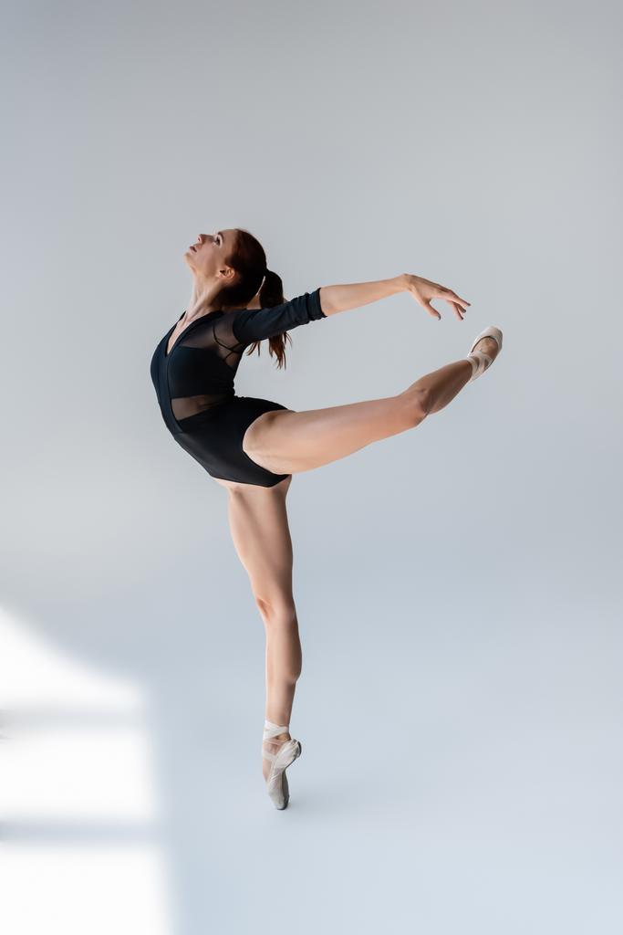 full length of flexible and young ballerina in black bodysuit dancing on grey - Photo, Image