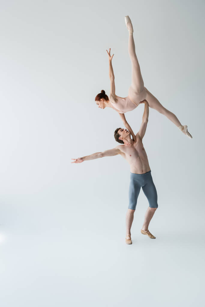 полная длина танцовщицы без рубашки, поднимающей изящную балерину в телесном костюме на сером - Фото, изображение