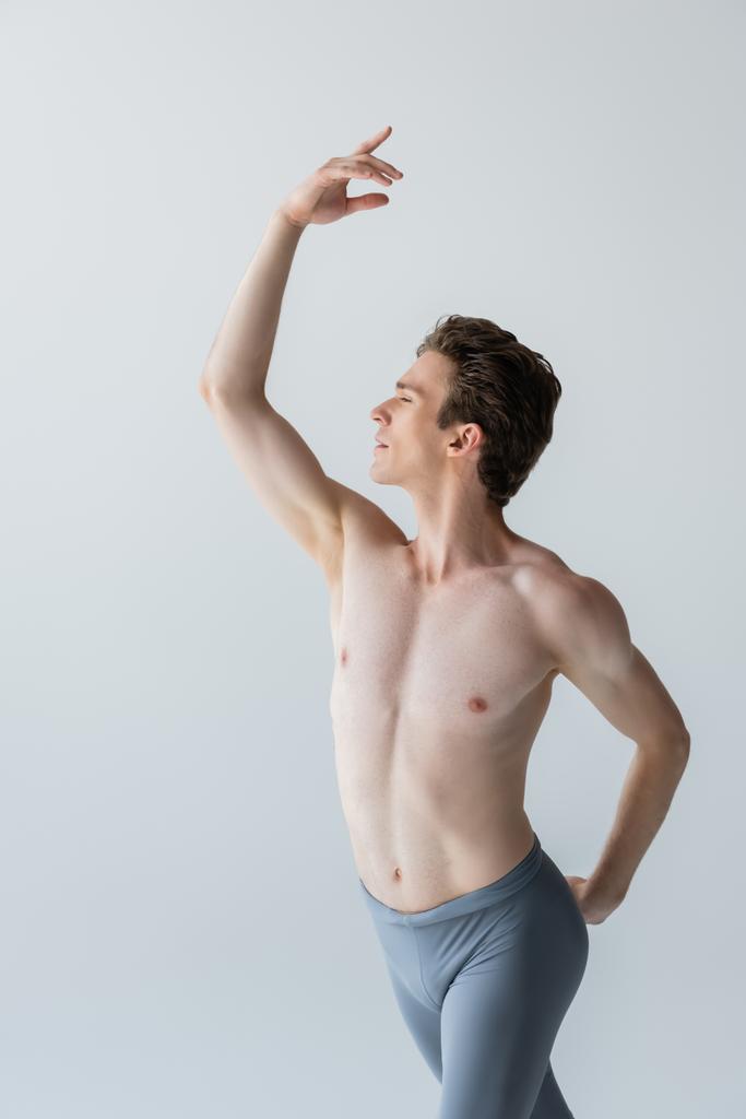 Mladý muž bez košile se zvednutou rukou provádějící baletní tanec izolovaný na šedi - Fotografie, Obrázek