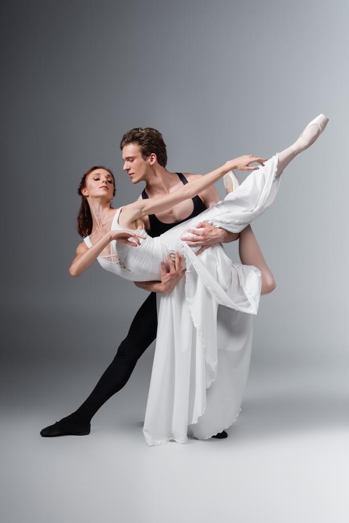 πλήρες μήκος του ανθρώπου υποστήριξη χαριτωμένη μπαλαρίνα σε λευκό φόρεμα, ενώ χορεύουν σε σκούρο γκρι - Φωτογραφία, εικόνα