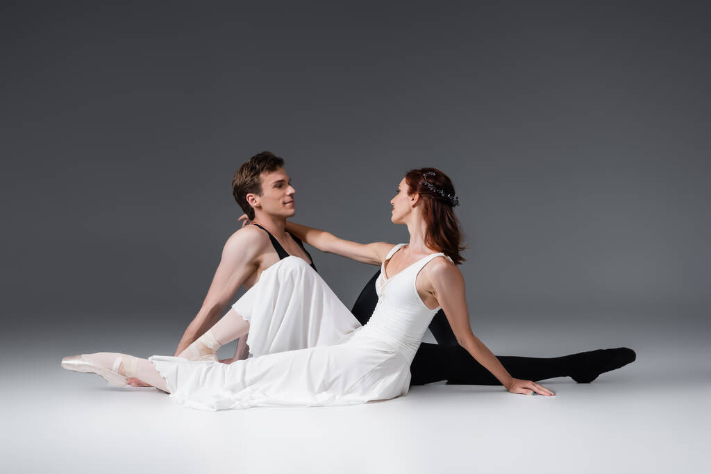 volledige lengte van sierlijke ballerina in witte jurk zitten met lachende partner op donkergrijs - Foto, afbeelding