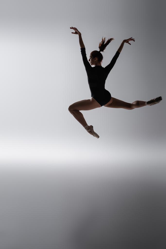 Рыжая балерина в боди-костюме прыгает с протянутыми руками на темно-сером - Фото, изображение