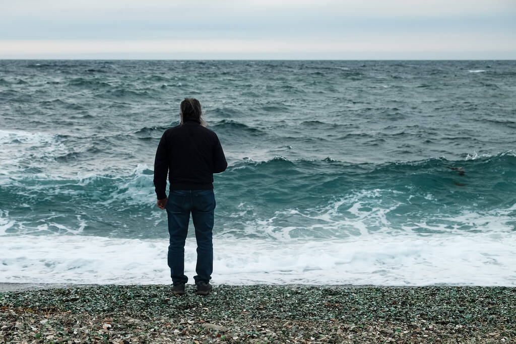 ένας άντρας στέκεται δίπλα στη θάλασσα μια συννεφιασμένη μέρα και κοιτάζει τα μεγάλα κύματα. - Φωτογραφία, εικόνα