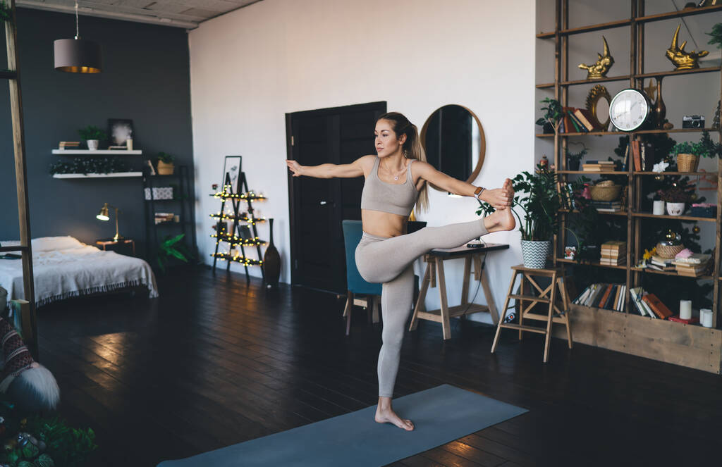 Європейська дівчина практикує йогу на фітнес маті вдома. Концепція здорового способу життя. Молода приваблива спортивна жінка стоїть на одній нозі. Внутрішня частина студії - Фото, зображення