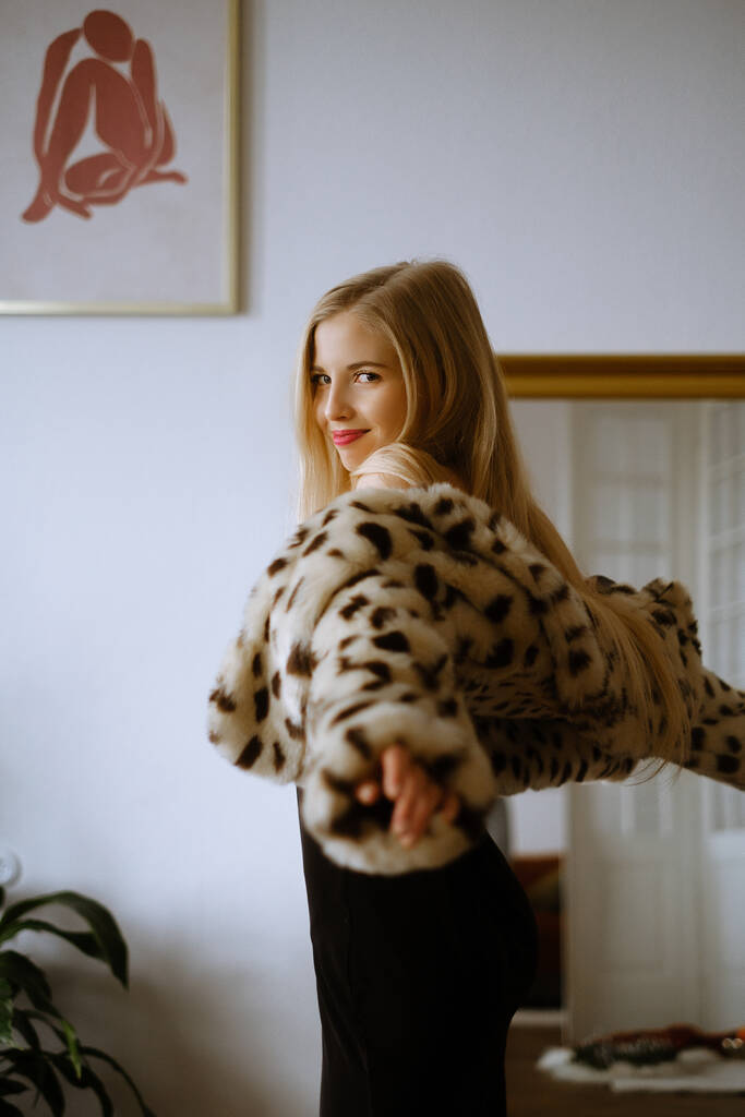 Porträt einer jungen kaukasischen Frau mit langen blonden Haaren, die einen Leopardenpelzmantel trägt, eines schönen schlanken Mädchens in einem Kleid und einem Pelzmantel in einer stilvollen Wohnung, einer hellen Dame mit Make-up in einem New Year - Foto, Bild