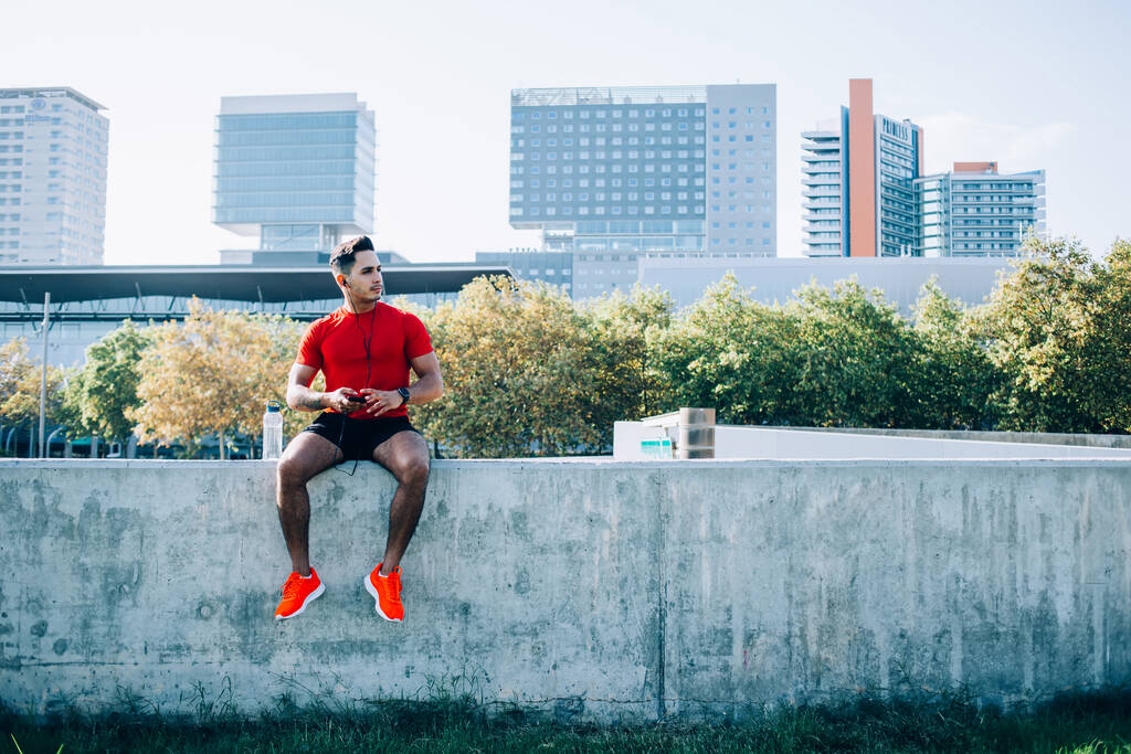 Serious ethnic young man in sportswear with earphones preparing for jogging using smartphone for listening music sitting on stone wall in stadium of modern city - Photo, Image
