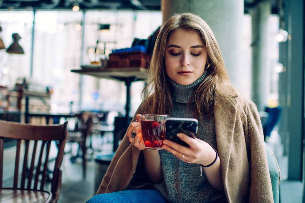 Mujer seria en traje elegante sentado en la cafetería moderna y el teléfono inteligente de navegación mientras bebe una taza de té caliente durante el fin de semana - Foto, Imagen