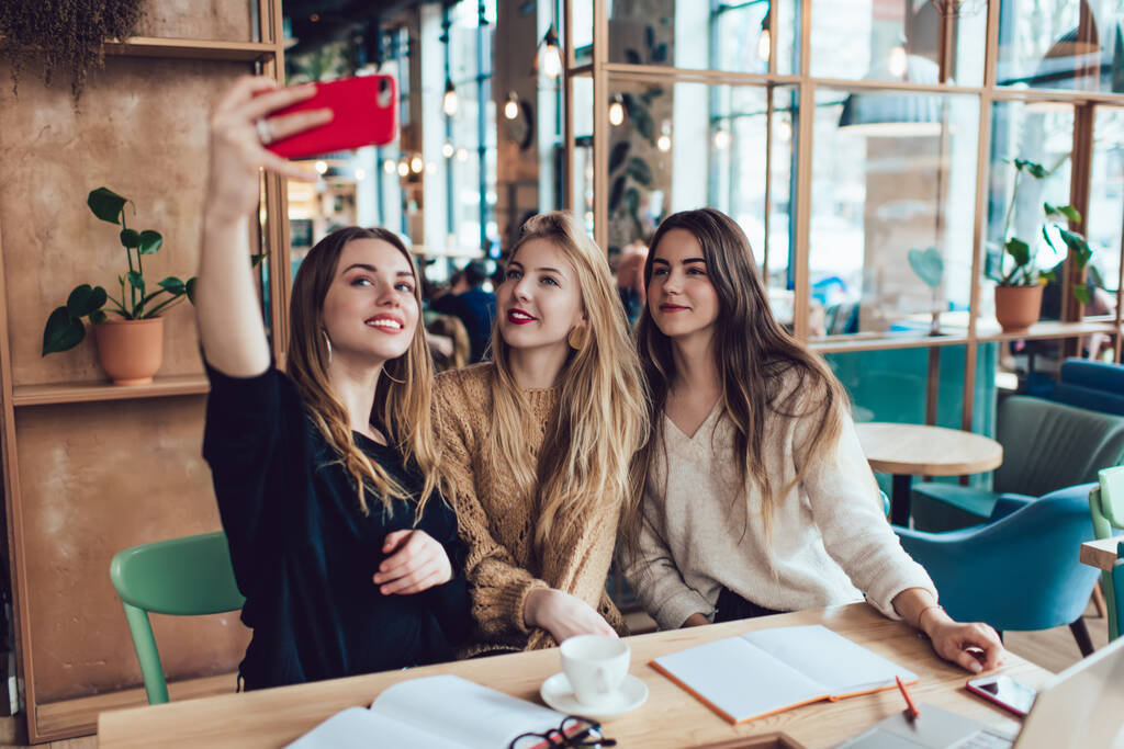 Mladé veselé ženy v ležérní oblečení přičemž selfie a při pohledu na kameru smartphone zatímco dělá domácí úkol společně v jasné anti kavárně - Fotografie, Obrázek