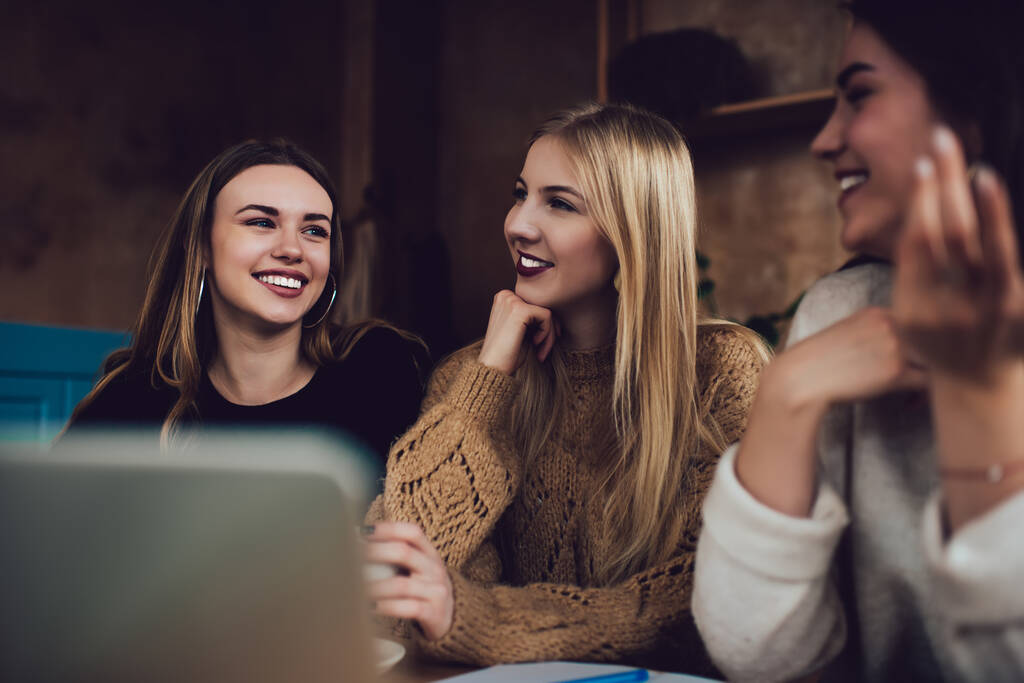 Jeunes étudiantes en vêtements décontractés se reposant et se regardant avec des sourires brillants tout en faisant des devoirs sur ordinateur portable depuis le café de temps - Photo, image