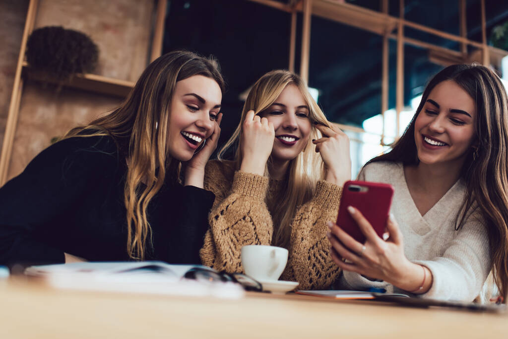 Kolme nuorta naista rento vaatteet älypuhelimella videoiden katseluun ja nauraa tehdessään läksyjä yhdessä ajoissa kahvilassa - Valokuva, kuva