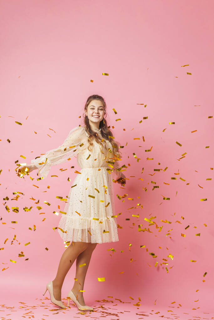 Buon Natale. Ciao. Una bella ragazza attraente con i capelli ondulati in un abito luminoso danza sotto la caduta d'oro, volando coriandoli su uno sfondo rosa in studio. In piena crescita. Copia spazio - Foto, immagini