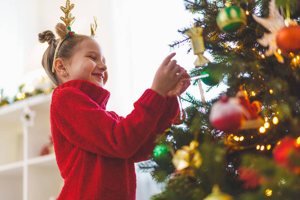 милая маленькая девочка украшая елку дома. Счастливого Рождества. Ребенок в красной куртке вешает игрушку на елку. - Фото, изображение