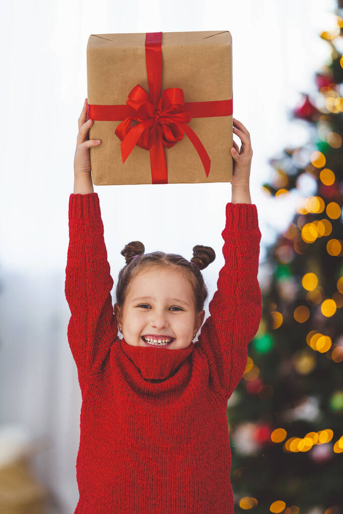 een schattig vrolijk klein meisje met een doos in haar handen, vastgebonden met een rood lint. Het kind glimlacht en houdt een geschenk boven zijn hoofd. Kerstboom in het licht van bloemenslingers op de achtergrond. - Foto, afbeelding