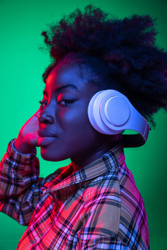 Muchacha joven de cerca, estudiante escuchando música en auriculares aislados sobre fondo de estudio verde oscuro en luz de neón púrpura. - Foto, imagen