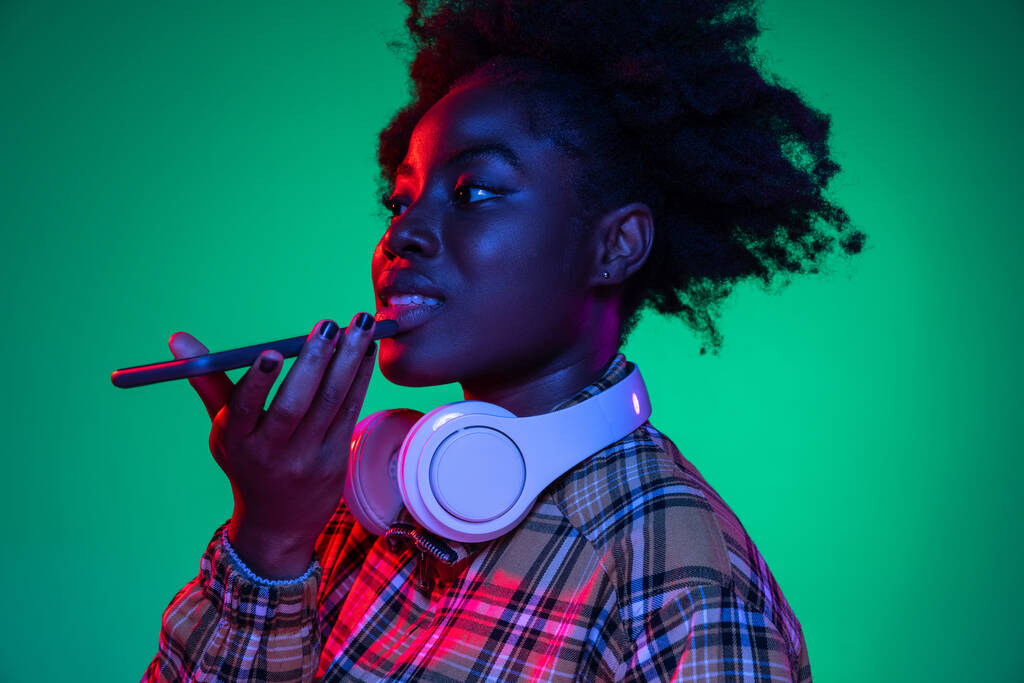 Studio záběr mladé krásné africké dívky, student pomocí telefonu izolované na tmavozeleném pozadí studia v purpurovém neonovém světle. - Fotografie, Obrázek