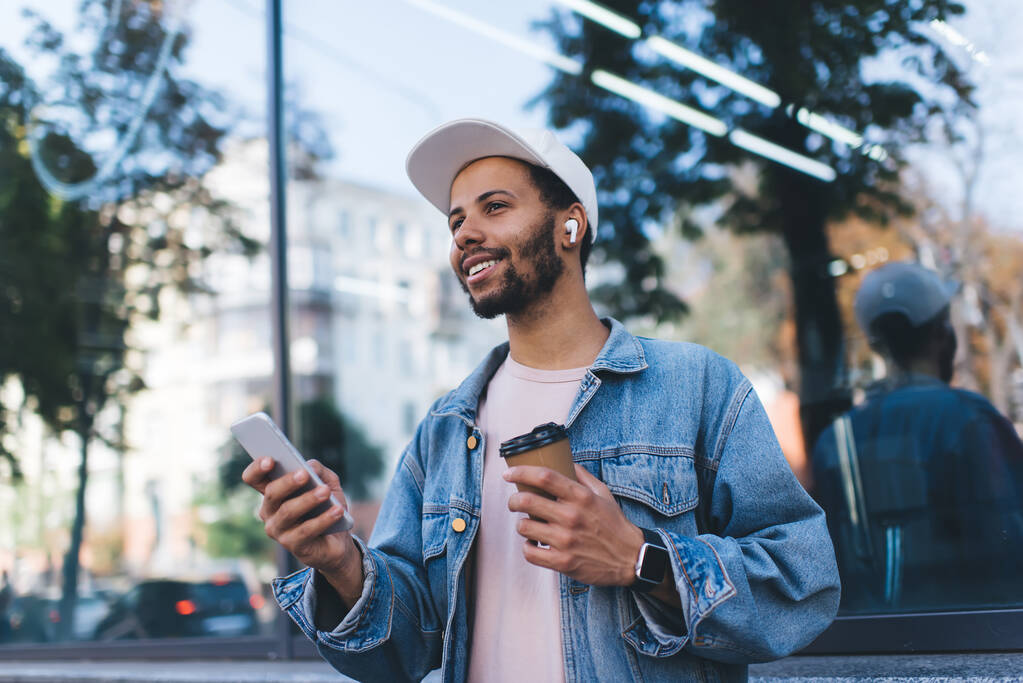 Улыбающийся испанский хипстер в наушниках Bluetooth наслаждается положительной музыкой плейлиста от мобильного радио с использованием 4g беспроводной в городе, тысячелетний турист в наушниках держит кофе, чтобы пойти и смартфон - Фото, изображение