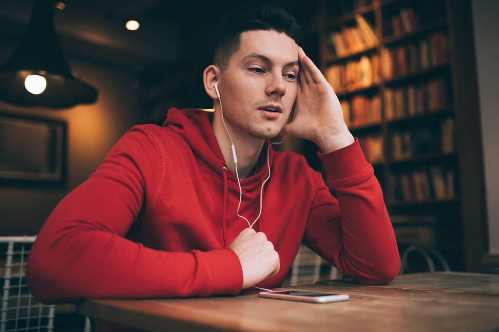 Hombre cansado con auriculares de mal humor sentado con la mano en la cabeza mientras escucha música y mira hacia otro lado pensativamente - Foto, Imagen