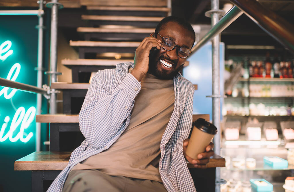 Χαρούμενος άνδρας σε οπτικά γυαλιά για να παρέχει διόρθωση όρασης γελώντας κατά τη διάρκεια της φιλικής συνομιλίας κινητών τηλεφώνων, αστείος μελαχρινός άνδρας με καφέ για να πάει να χαρεί κατά τη διάρκεια της κινητής ομιλίας - Φωτογραφία, εικόνα