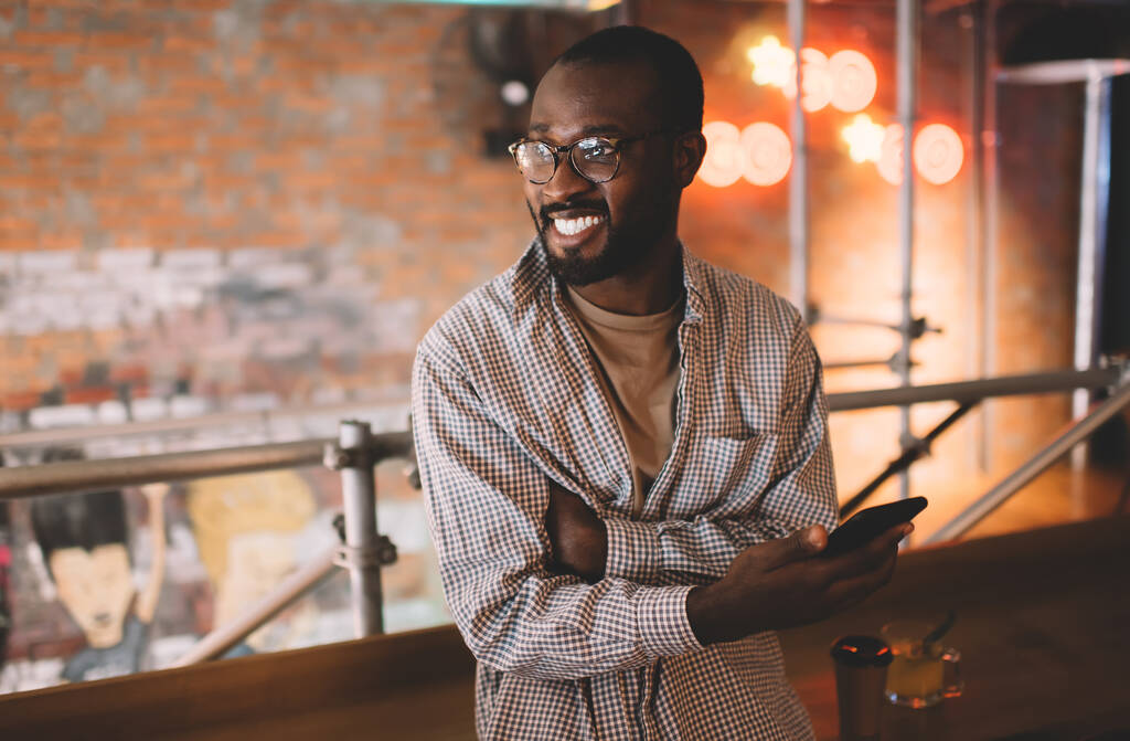 Χαρούμενος hipster τύπος σε κομψά γυαλιά χρησιμοποιώντας κινητό gadget για την κοινωνικοποίηση του δικτύου κατά τη διάρκεια του ελεύθερου χρόνου, ευτυχισμένος Αφροαμερικανός άνδρας σε οπτικά γυαλιά κρατώντας συσκευή smartphone και χαμογελώντας - Φωτογραφία, εικόνα